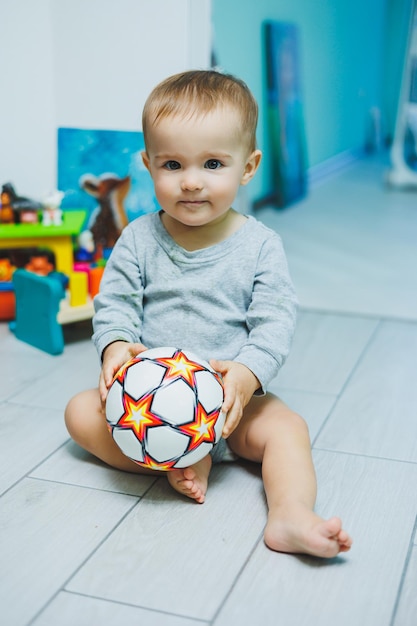 A little blond boy is sitting on the floor in a room with bare heels of his legs making grimaces and holding an orange balloon in his hands