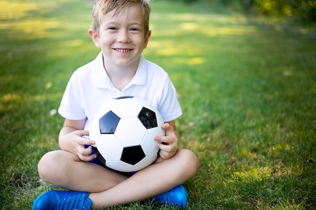 어린 금발 소년이 여름에 카메라를 보며 축구공을 들고 잔디에 앉아 있다