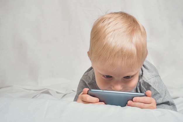 Little blond boy looking something his nose buried in a smartphone, lying in bed. Gadget Leisure