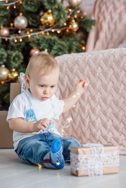 Little blond boy plays with a bright garland at the Christmas tree at cozy home.