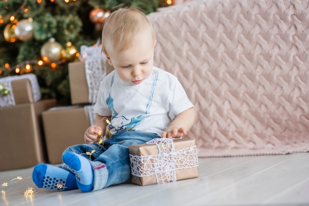 小さな金髪の少年は、居心地の良い家のクリスマスツリーで明るい花輪で遊んでいます。