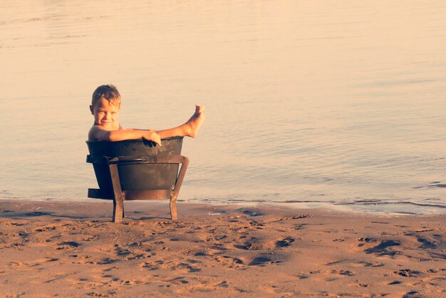 Little blond boy sitting in rusty pot on the beach in the sunlight