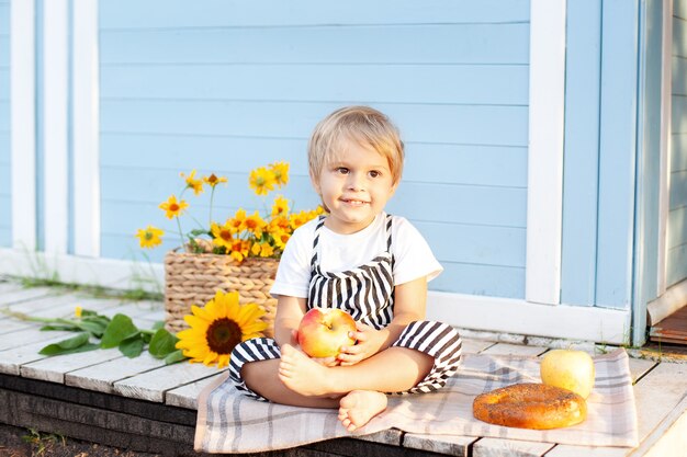 Little blond boy sitting on a wooden porch at home and eats an apple Happy child and Fruit