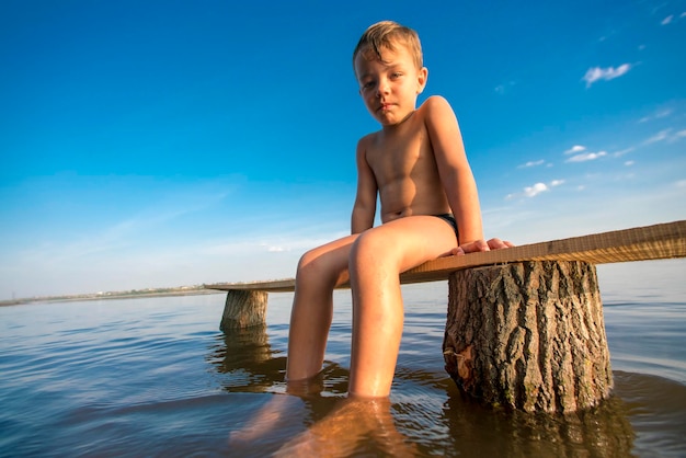水の中の木のベンチに座る水泳パンツを着た金髪の少年