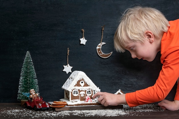 Little blond boy touches Christmas composition from gingerbread house, toy car and Christmas tree.