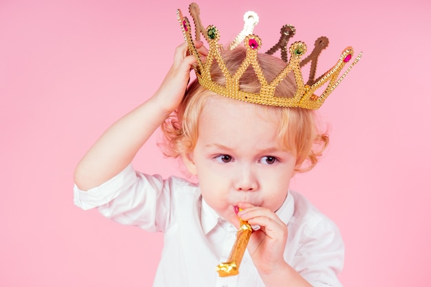 Little blond boy with golden crown head curls hairstyle 4-5 year old in studio on pink background blowing noisemakers horn-whistle a birthday party celebrates christmas and new year king of the party