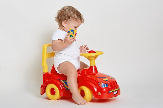 Little blond driver sitting on red tolokar, wearing white bodysuitac