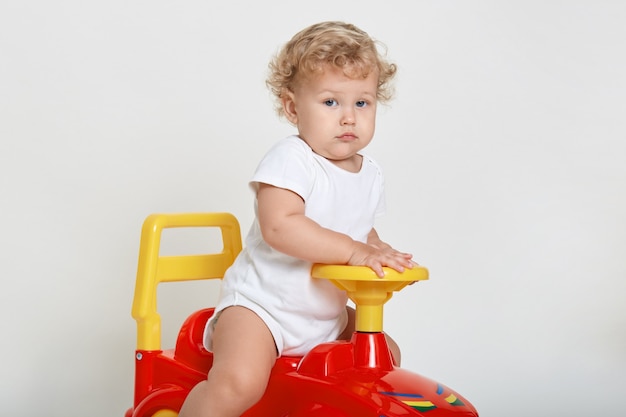 Little blond driver sitting on red tolokar, wearing white bodysuitac