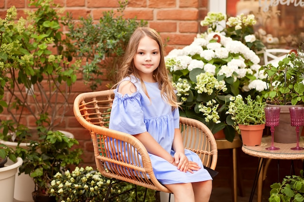 Little blond girl in blue dress sitting on the chair in the garden