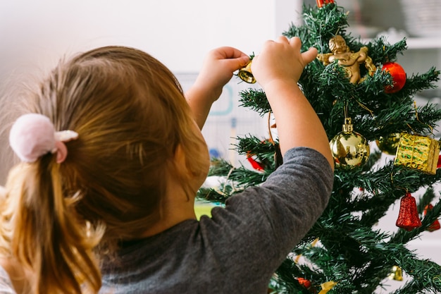 写真 背中を向けた小さなブロンドの女の子が家でクリスマスツリーを飾る