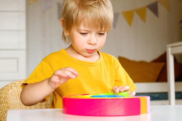 Little blond hair boy in clothes natural fabric plays rainbow colored wooden toys at white table Eco