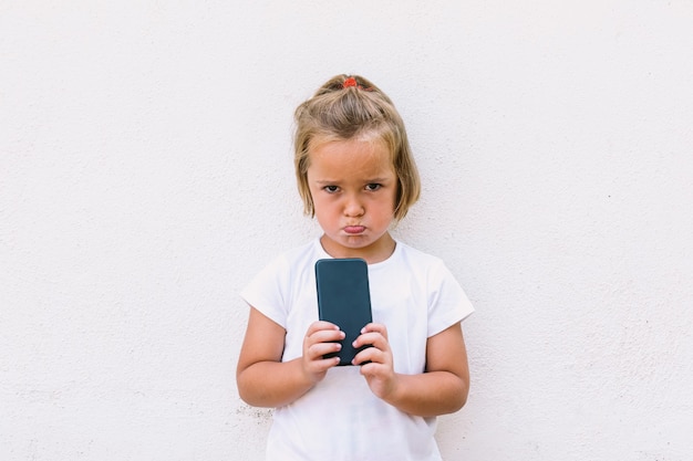 Little blond hair girl wearing white t-shirt, holding mobile phone, looking angry