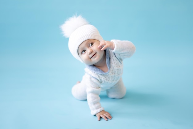 Little blonde baby girl in a white winter knitted hat and knitted sweater on a blue background isolate, space for text, baby toddler in winter clothes