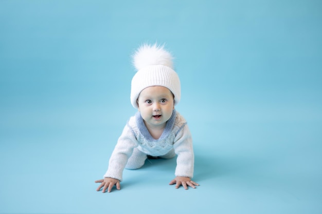 Little blonde baby girl in a white winter knitted hat and knitted sweater on a blue background isolate, space for text, baby toddler in winter clothes