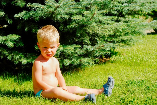 Little blonde boy sits on the grass in summer park