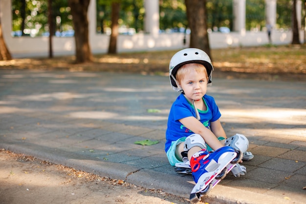 Little blonde boy in white sport helmet and blue t-shirt riding on the roller-skates