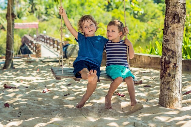 Little blonde boys sitting on a swing on a tropical beach