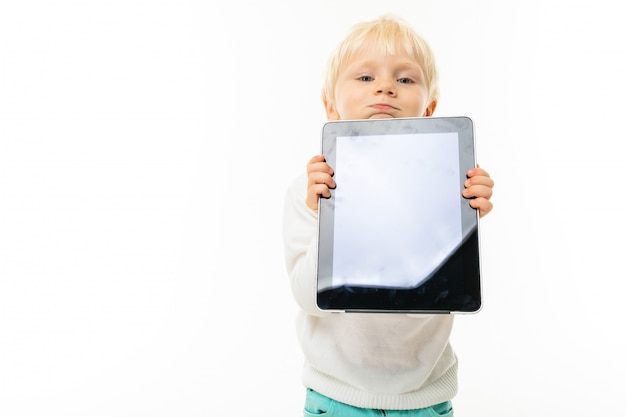 Little blonde caucasian boy shows the tablet isolated on white wall