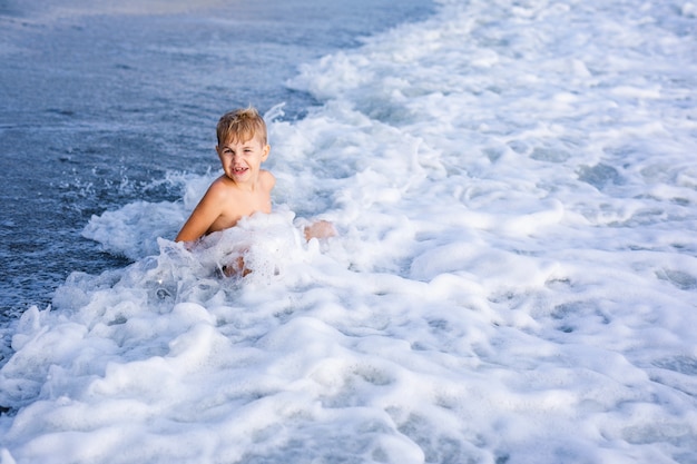 Little blonde child boy siting on sea shore and playing in waves