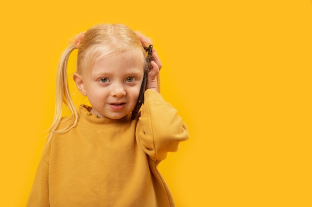 Photo little blonde girl 45 years old wearing yellow sweatshirt use mobile cell phone studio portrait of kid isolated on yellow background