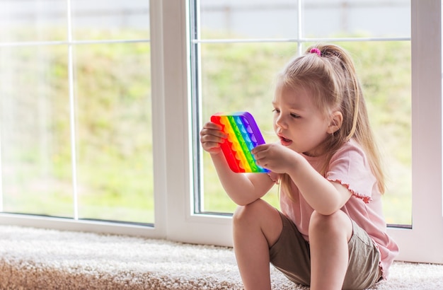 小さなブロンドの女の子の子供たちは、新しいトレンドの感覚的なおもちゃの虹で遊んでいます。子供と大人のための抗ストレスおもちゃ。カラフルなおもちゃのシンプルなディンプル。スクイーズソフトバブルおもちゃレインボーカラー