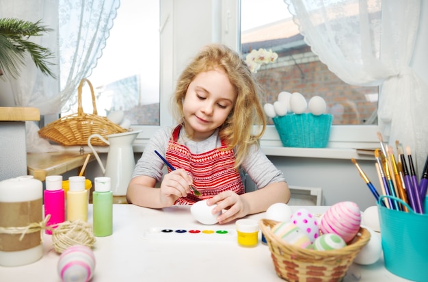 Little blonde girl coloring eggs for Easter holiday at home.