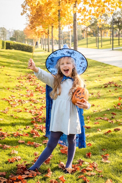 魔女、ハロウィーンの魔法使いに扮した小さなブロンドの女の子。帽子、マント、カボチャ、笑い、笑顔、秋の休日