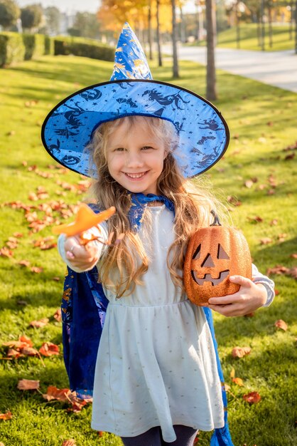 魔女、ハロウィーンの魔法使いに扮した小さなブロンドの女の子。帽子、マント、カボチャ、笑い、笑顔、秋の休日