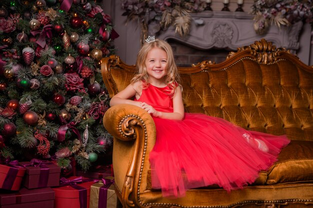 little blonde girl in evening red dress near the Christmas tree