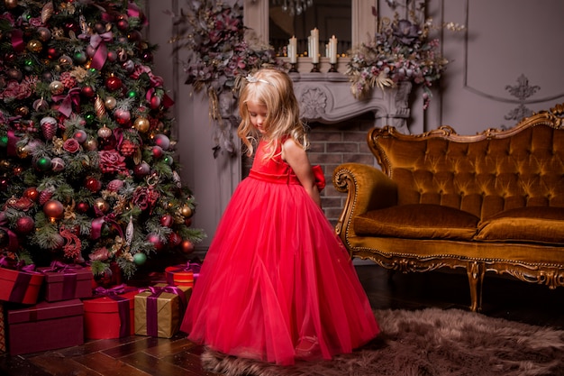 little blonde girl in evening red dress near the Christmas tree