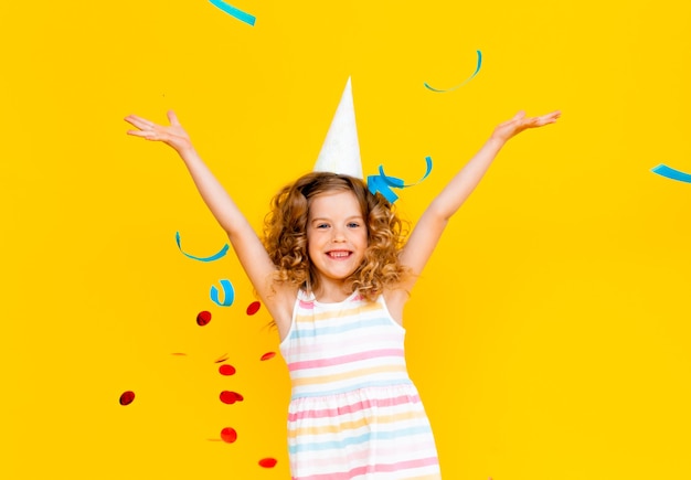 Little blonde girl in a festive cap and in a white dress catches confetti a happy smile on a yellow background.