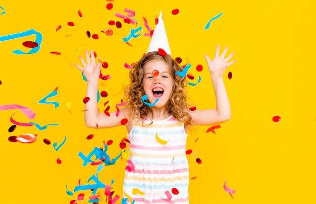 Photo little blonde girl in a festive cap and in a white dress catches confetti a happy smile on a yellow background.