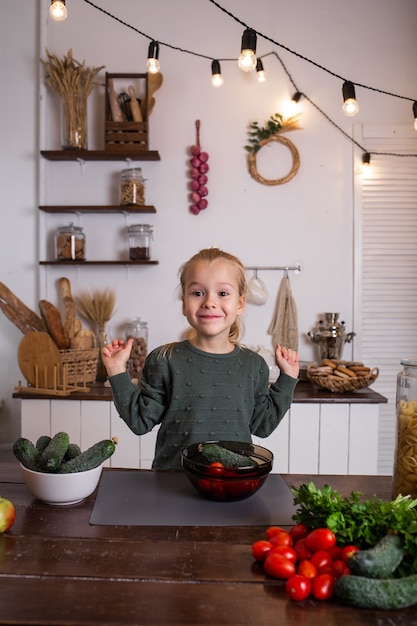 A little blonde girl in a green jumper is sitting at a wooden table and preparing a vegetable salad