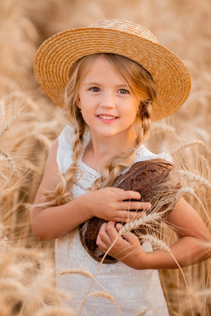 little blonde girl holds a fresh loaf of bread in a wheat field in the summer