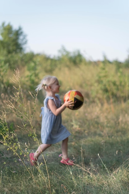 Фото Маленькая блондинка в сарафане бежит с мячом в руках по полю ребенок играет с мячом фото в движении вертикальная рамка