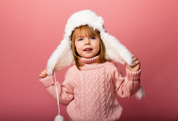 Маленькая блондинка в зимней одежде и меховой шапке на розовом фоне.