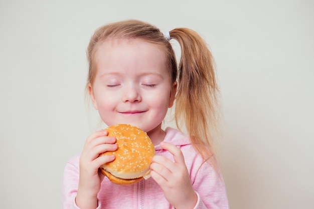 小さなブロンドの女の子は野菜と一緒に焼きたてのベジタリアンハンバーガーを食べています。子供のビーガンのアイデア健康的な食事の概念。