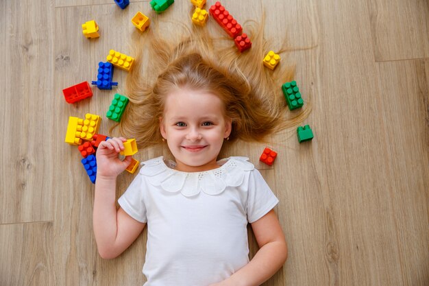 A little blonde girl is lying on the floor among the constructor. The view from the top.