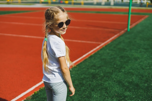 小さなブロンドの女の子がスポーツフィールドで走っていて、彼女はサングラスと白いTシャツを着て笑っています夏休みと休暇の時間