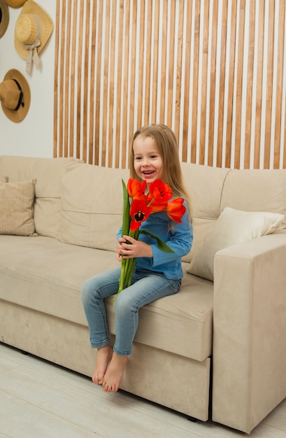 Una bambina bionda è seduta sul divano e tiene in mano i tulipani rossi