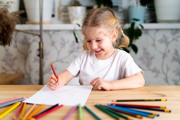 A little blonde girl is sitting at the table smiling and drawing a heart with a red pencil there are