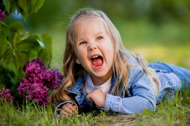 Little blonde girl lies near the lilac flowers on the green grass and smiles