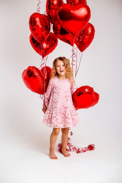 маленькая белокурая девушка в розовом платье улыбается и держит много красных сердцевидных воздушных шаров
