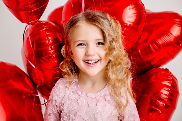 Bambina bionda in un abito rosa è sorridente e in possesso di un sacco di palloncini a forma di cuore rosso su un muro bianco concetto di san valentino