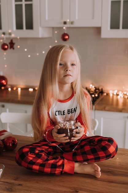 빨간 크리스마스 잠옷을 입은 금발 소녀가 장식된 부엌에서 뜨거운 음료와 마시멜로가 든 유리잔을 들고 있습니다. 겨울 주말 및 공휴일. 생활 양식. 텍스트를 위한 공간입니다. 고품질 사진