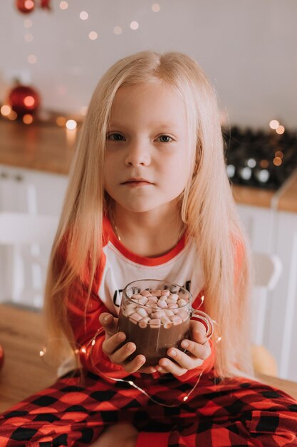 Little blonde girl in red christmas pajamas holding a glass mug with a hot drink and marshmallows in a decorated kitchen. winter weekends and holidays. lifestyle. space for text. High quality photo