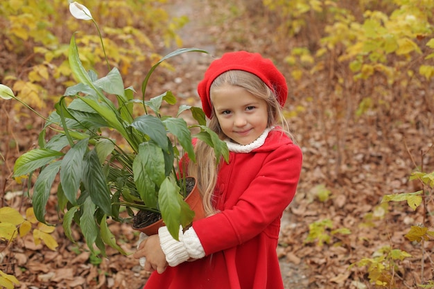 赤いコートとベレー帽をかぶった小さなブロンドの女の子は、路上で彼女の手に鉢植えの花を持っています