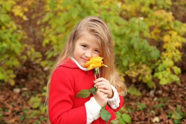a little blonde girl in a red coat holds a yellow rose in her hands in the park
