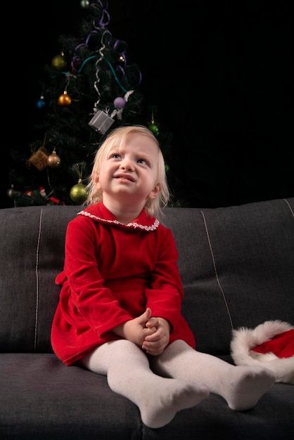 Little blonde girl in a red dress sits on the sofa and dreams against the background of a Christmas tree and a black wall Christmas Eve New Year