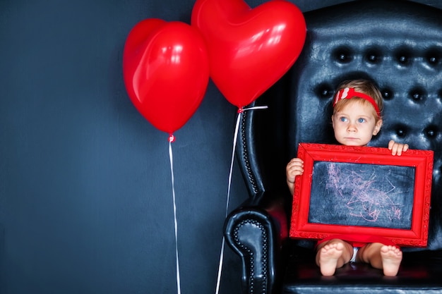 Little blonde girl in red dress with red wreath with hearts sitting on the armchair with red heart balloon on the St. Valentine's day. 